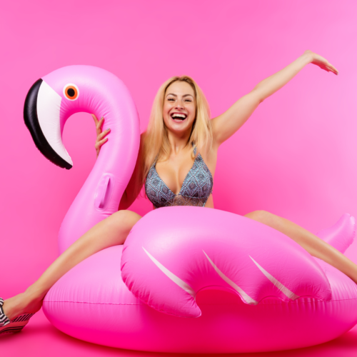 Glad ljushyad och blond kvinna som håller upp en hand i luften. Kvinnan är iklädd grå bikini och sittandes på uppblåsbar flamingo mot rosa bakgrund. Foto.